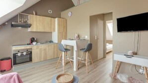 A modern kitchen with wooden cabinets, a dining area with a table and chairs, a television mounted on the wall, and a cozy seating area with a basket and a fluffy rug. - Varde - appartement avec jardin (Saint-Coulomb)