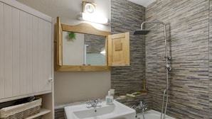Een badkamer met een wit wastafelmeubel, houten kastjes, een spiegel, een douche en een klok aan de muur.