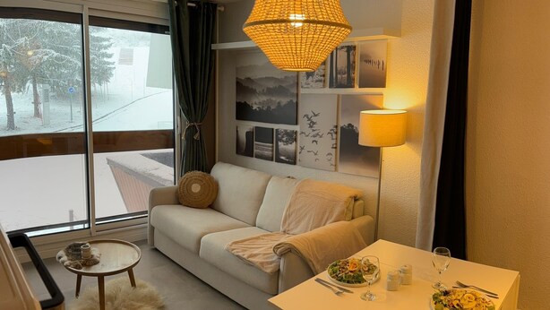 Een gezellige woonkamer met een bank, een eettafel gedekt met eten, een lamp en een groot raam met uitzicht op een besneeuwde omgeving.