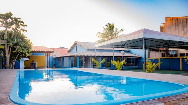 A swimming pool with a tiled deck, surrounded by buildings with blue and yellow exteriors, and a covered area with a roof. - Ribeiro Lugar de Paz Casa 1 (Arraial do Cabo)
