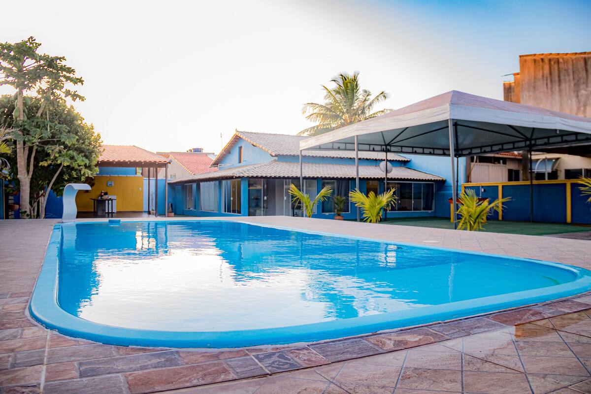 A swimming pool with a tiled deck, surrounded by buildings with blue and yellow exteriors, and a covered area with a roof.