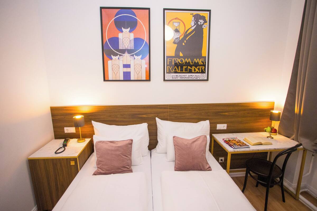 Une chambre d’hôtel avec deux lits, une tête de lit en bois, des lampes de chevet, une petite table avec un livre et une lampe, et deux œuvres d’art encadrées accrochées au mur.