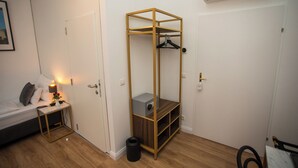 A compact room with a bed, bedside table, lamp, and a wooden storage unit with shelves and a hanging rack. - Pension Central - Self Check-in (Vienna)
