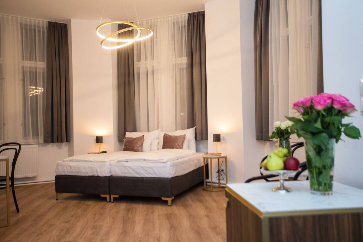 Une chambre moderne avec un lit double, deux tables de chevet avec des lampes, une plante sur l’une des tables de chevet et une vue sur la ville à travers des rideaux transparents.
