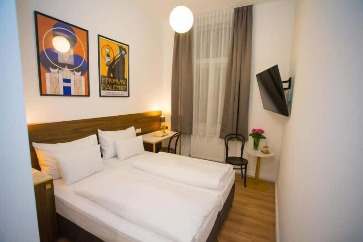 Une chambre d’hôtel avec un lit double, une petite table, une chaise, un téléviseur fixé au mur et deux œuvres d’art encadrées.