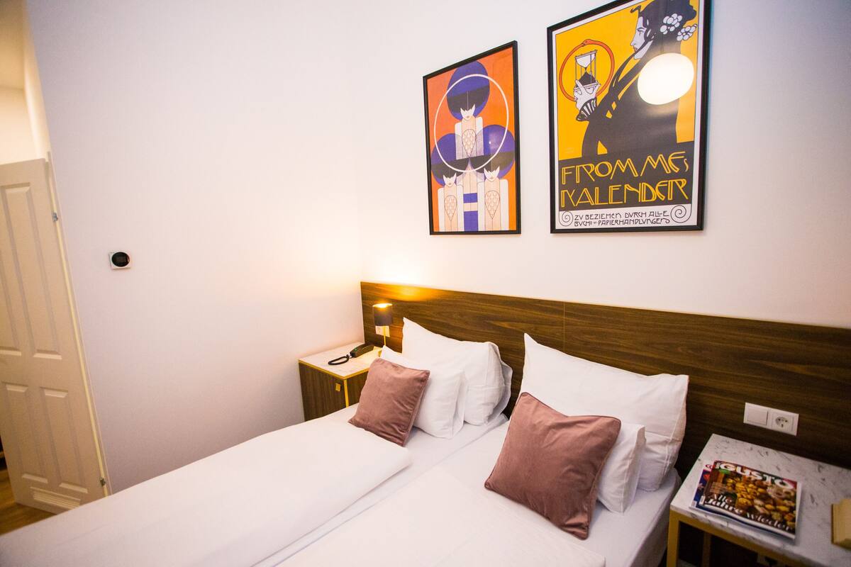 Un lit bien fait, avec des oreillers blancs et bruns, une tête de lit en bois, une table de chevet avec une lampe et deux affiches encadrées au mur.