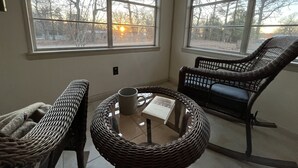 Una habitación con ventanas amplias, mobiliario de mimbre y una mesa con un libro y una taza.