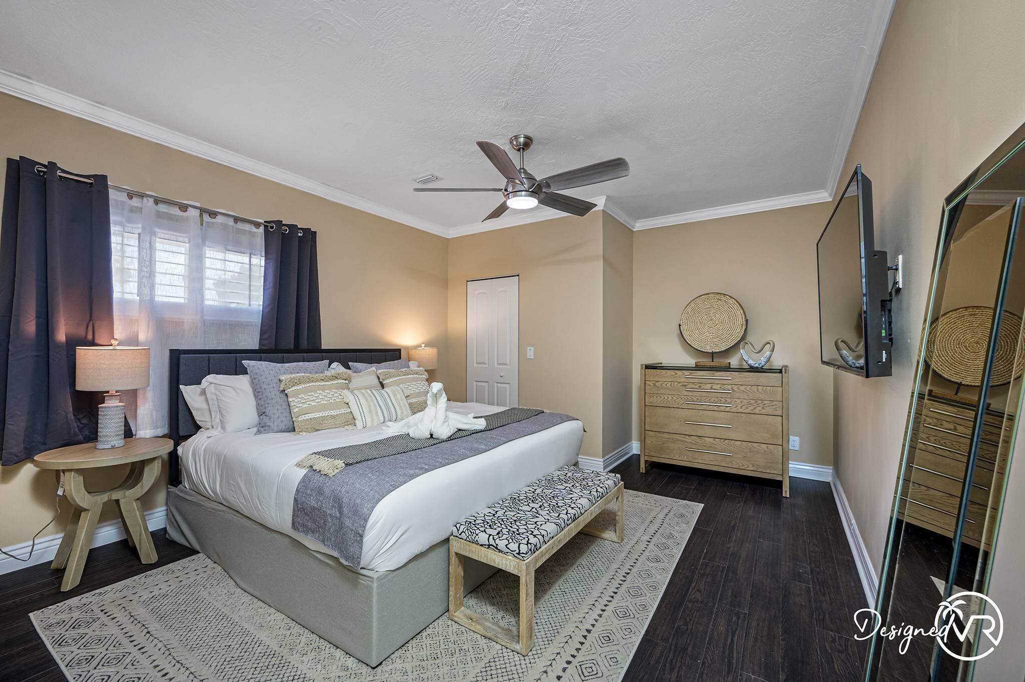 Een slaapkamer met een bed, nachtkastjes, een plafondventilator, een dressoir, een spiegel en een raam met gordijnen.