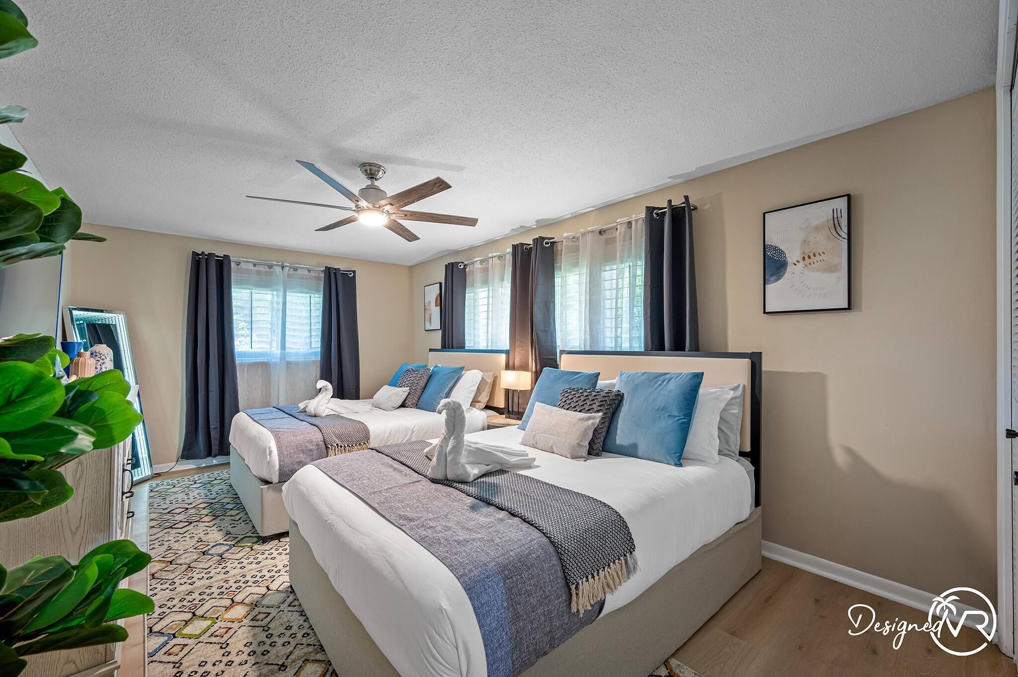 A hotel room with two beds, a ceiling fan, a large window with curtains, a framed picture on the wall, and a patterned rug.