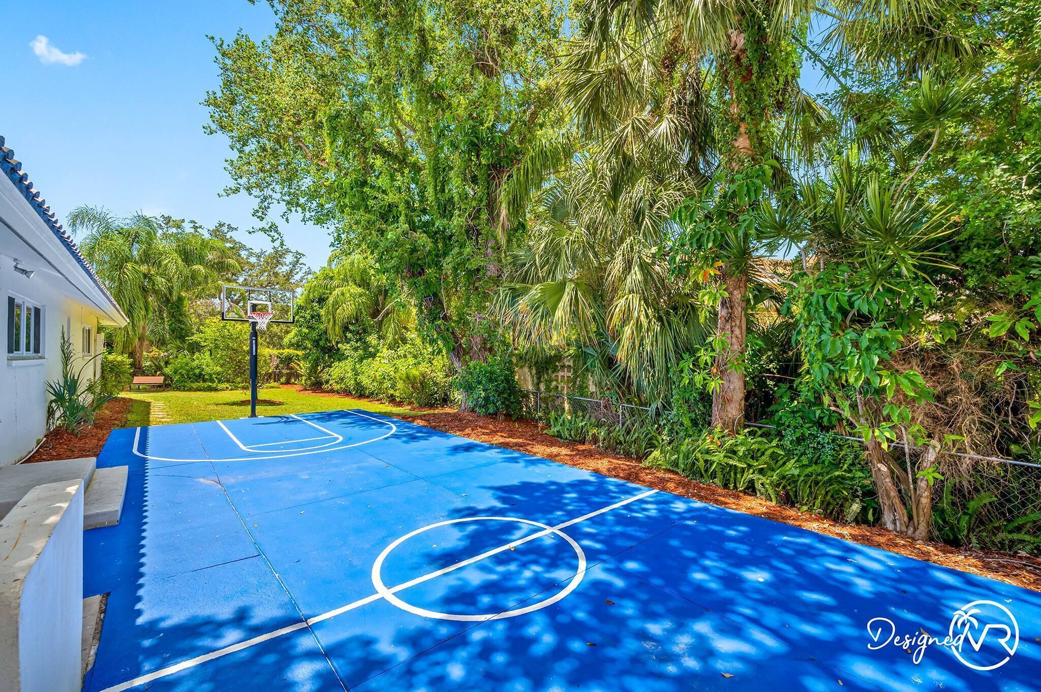 An outdoor basketball court with a blue surface, surrounded by lush greenery and a white building.