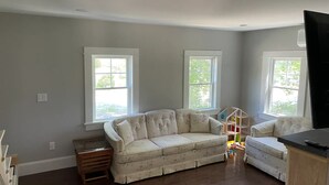 Un salón con un sofá color beige, dos ventanas, una mesita de madera y un televisor.