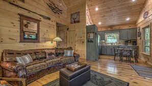 A log cabin interior with a wooden ceiling, a sofa with animal prints, a dining table with chairs, and a kitchen area. - Relaxing Getaway! Cozy Log Cabin, Hot Tub + Firepit (Cherry Log)