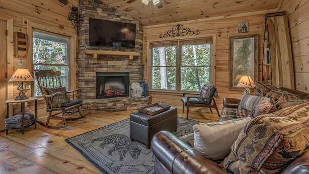 A cozy cabin interior with a stone fireplace, wooden floors, and a large window. - Relaxing Getaway! Cozy Log Cabin, Hot Tub + Firepit (Cherry Log)