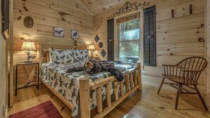 A wooden cabin bedroom with a bed, two bedside lamps, a chair, and a window with shutters. - Relaxing Getaway! Cozy Log Cabin, Hot Tub + Firepit (Cherry Log)