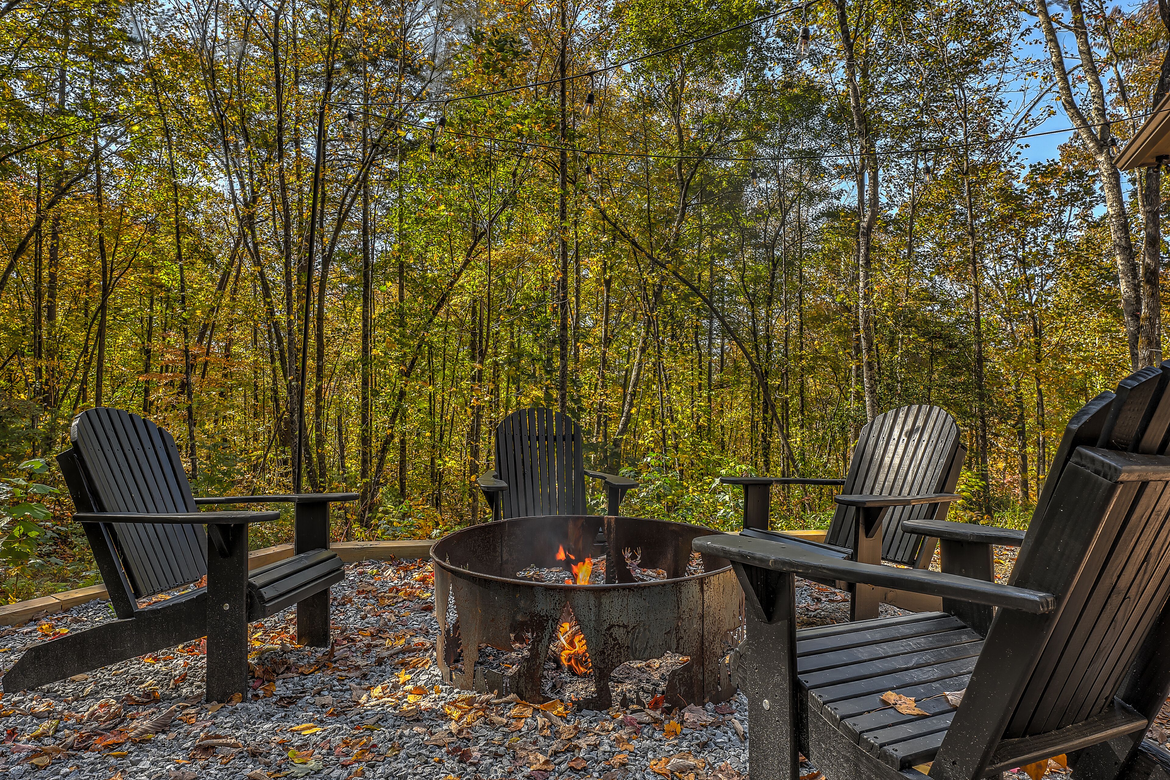 A circular fire pit with three Adirondack chairs around it, set on a gravel surface with fallen leaves.