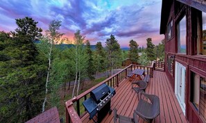 A wooden deck with a grill, outdoor seating, and a view of a forested landscape. - Window of the Rockies! (Bailey)