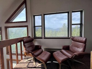 A room with a view of mountains through large windows, a wooden railing, and two leather chairs. - Window of the Rockies! (Bailey)