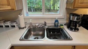 A kitchen with wooden cabinets, a double sink, a coffee maker, and a window with a view of trees. - Cozy Retreat in the Woods & Gateway to Adventure (Skandia)
