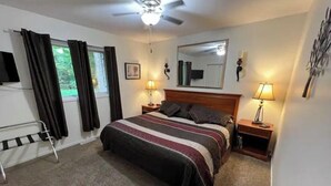 A bedroom with a bed, two bedside lamps, a ceiling fan, a window with curtains, a bedside table, and a mirror on the wall. - Cozy Retreat in the Woods & Gateway to Adventure (Skandia)