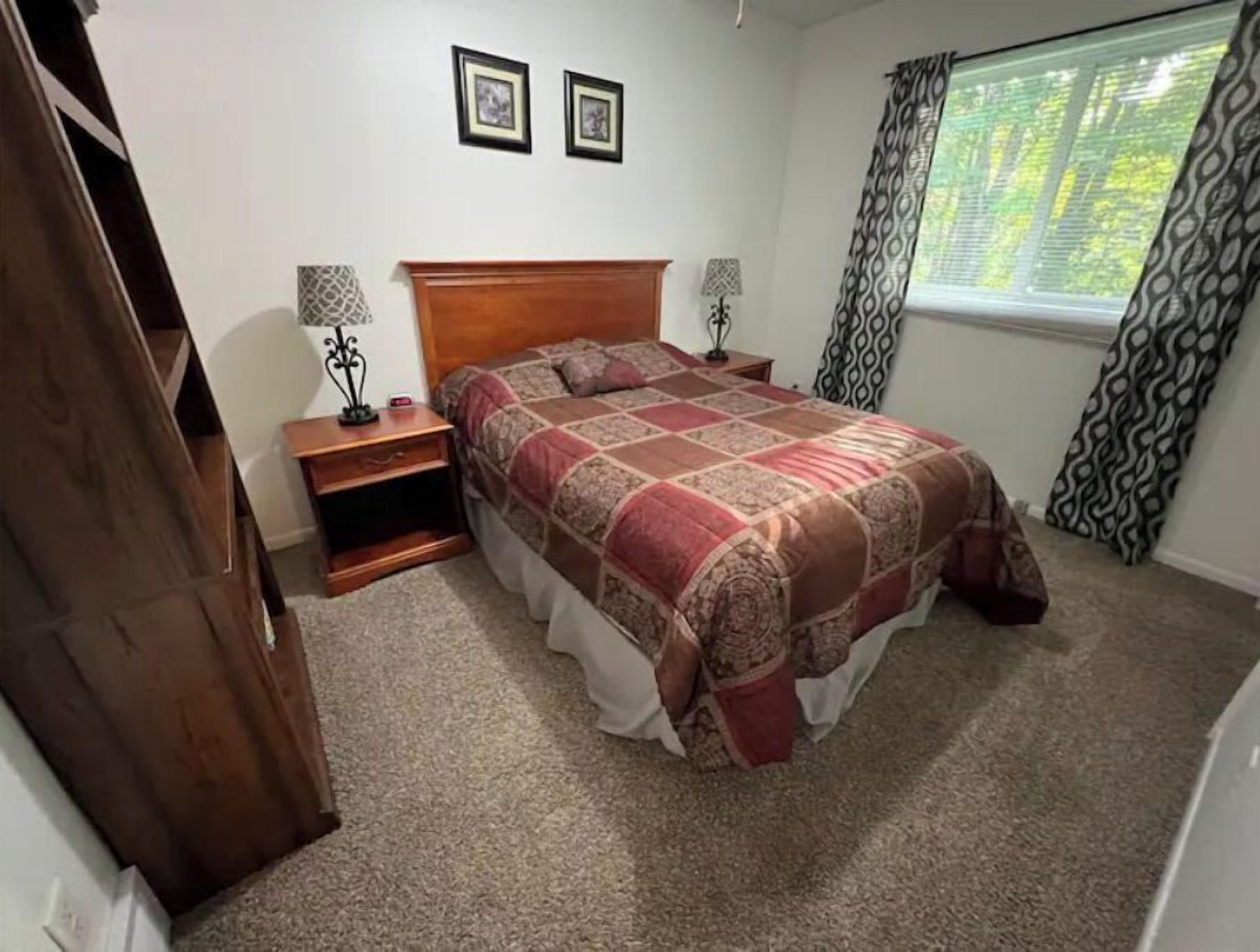 A bedroom with a wooden bed, two bedside tables, a nightstand, a patterned bedspread, and a window with curtains.