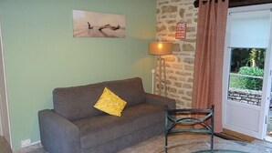 A room with a sofa, a lamp, a chair, a door with a view, and a stone wall.