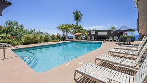 A rectangular pool with a clear blue waterline, surrounded by lounge chairs and a small table with umbrellas. - NEW! Quiet Ocean-view 1st floor | Patio | Pool (Kailua Kona)