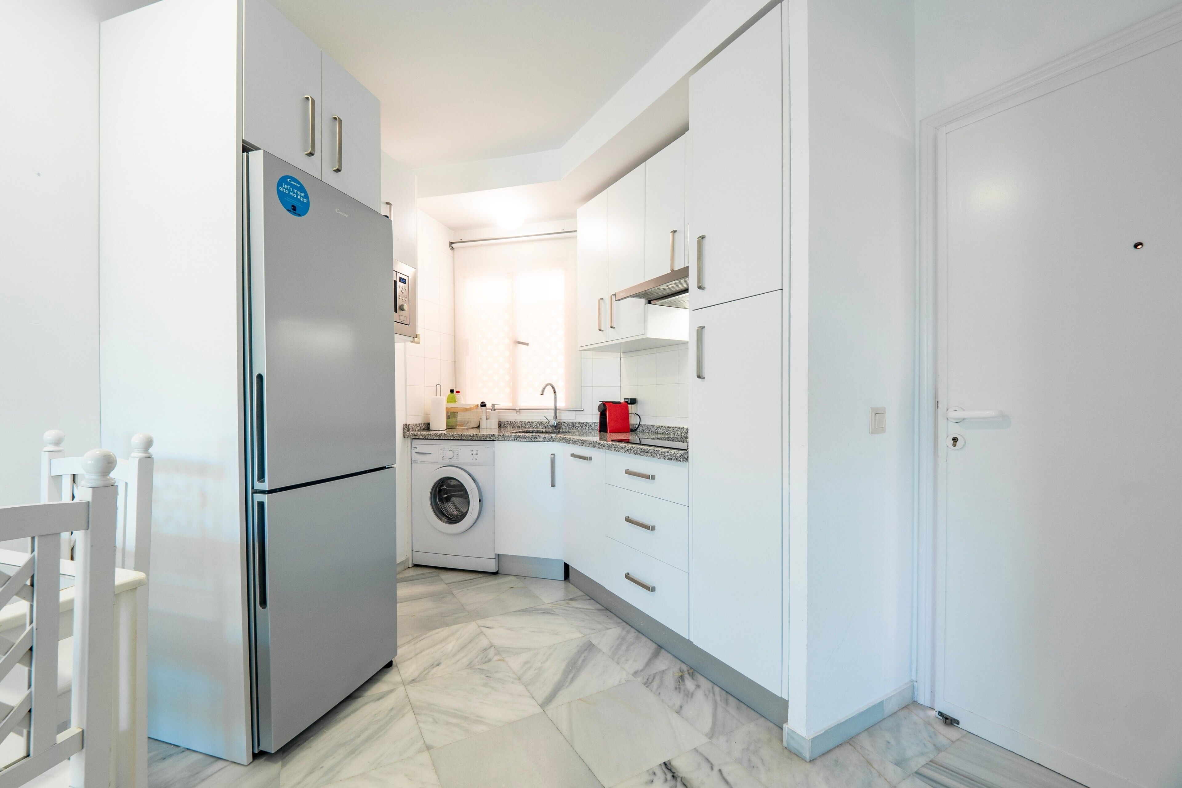 Una cocina moderna con armarios blancos, un refrigerador de acero inoxidable, una lavadora y un hervidor rojo.