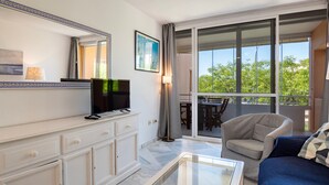 Ein modernes Hotelzimmer mit Flachbildfernseher, großem Fenster mit Blick auf Grünflächen, Balkon mit Esstisch und einer Couch.