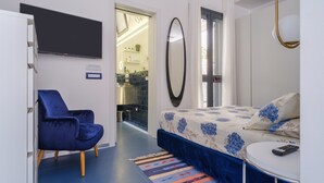 Un dormitorio con cama, una silla, un televisor, un espejo y un baño visible a través de una puerta abierta.