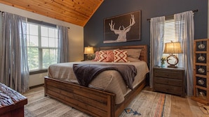 A bedroom with a wooden bed frame, a large window with curtains, a wall-mounted lamp, and a decorative deer head on the wall. - Fall beauty! Hot tub, Family Friendly, Games Firepit, Pool/Marina Access (Galena)