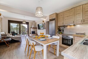 A modern kitchen with wooden cabinets, a dining area with a wooden table and chairs, a built-in oven, and a large window offering a view.