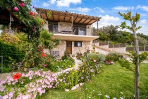 A two-story stone house with a balcony, surrounded by a well-maintained garden with various flowers and plants.