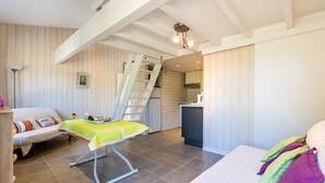 A bedroom with a bed, a chair, a desk, a ladder, and a kitchenette.