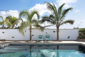 Une piscine avec deux chaises longues, des palmiers et un mur blanc.