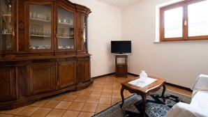 A living room with a wooden cabinet, a television on a stand, a white chair, and a patterned rug. - Holiday Home 'Tramonto sul Castello di Poppi' with View, Hot Tub, Garden & Wi-Fi (Poppi)