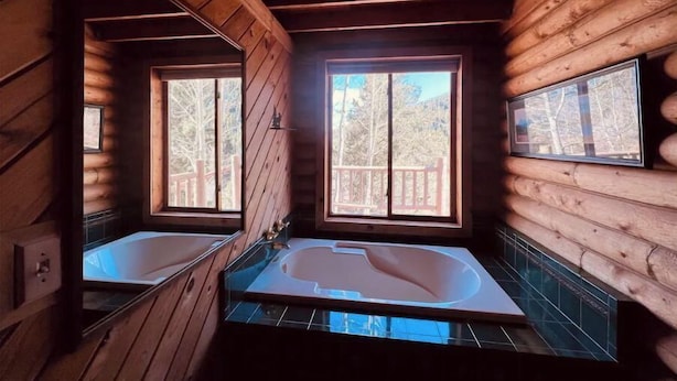 Un baño de cabaña con dos ventanas amplias, detalles de madera y una bañera de hidromasaje con mampara de vidrio.