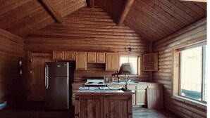 Interior de una cabaña de troncos con una zona de cocina que incluye armarios de madera, un refrigerador, una estufa y una ventana que deja entrar luz natural.
