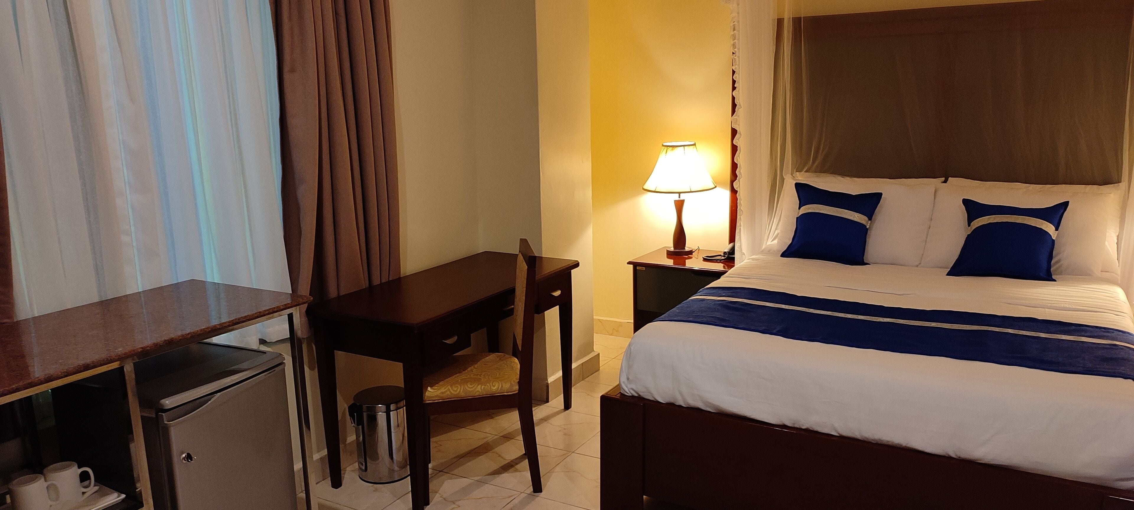 Une chambre d’hôtel comprenant un lit, un bureau, une chaise, une poubelle et une table de chevet avec une lampe.