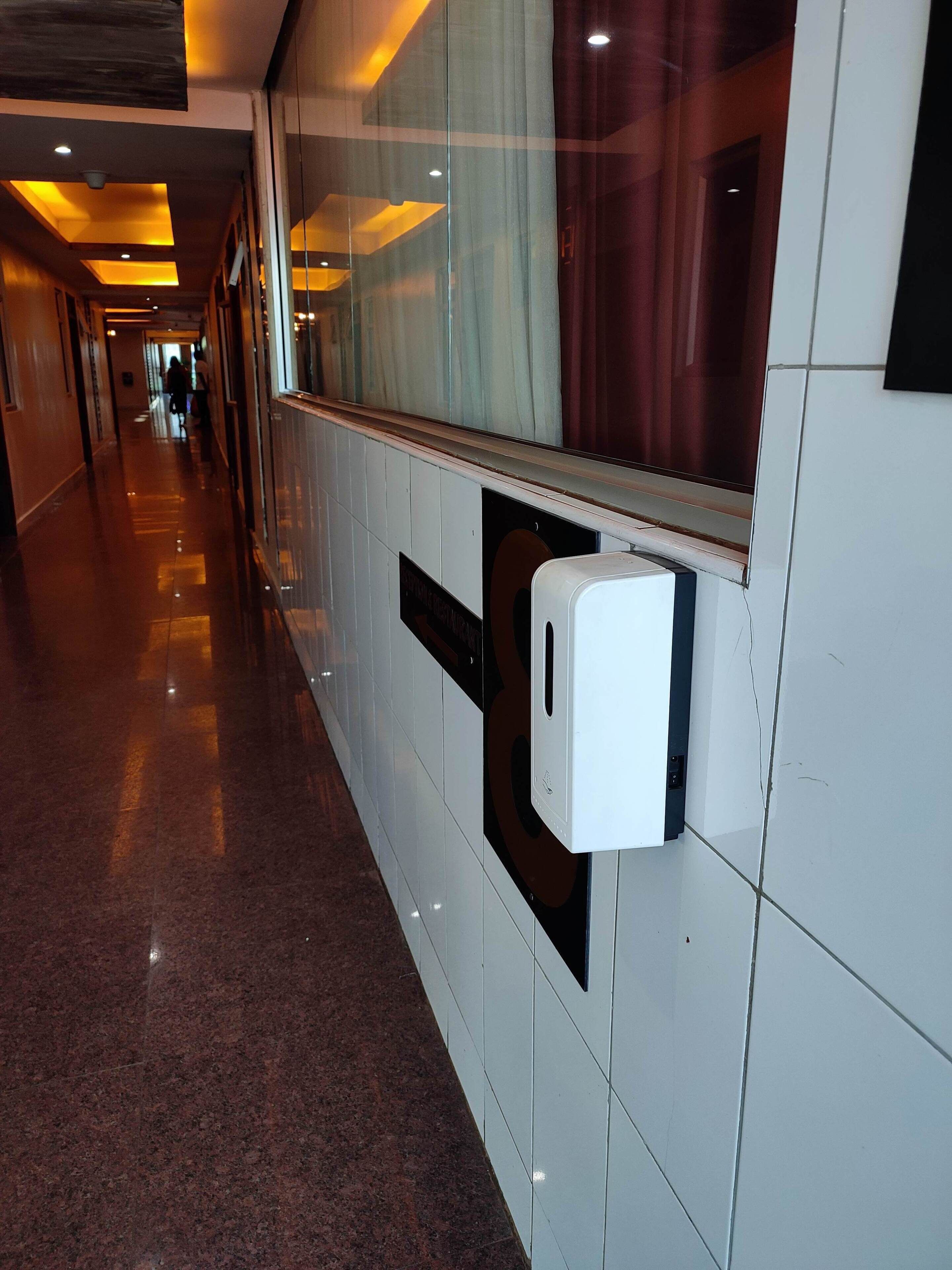 A corridor with a hand sanitizer dispenser mounted on the wall, a tiled floor, and a window with curtains.