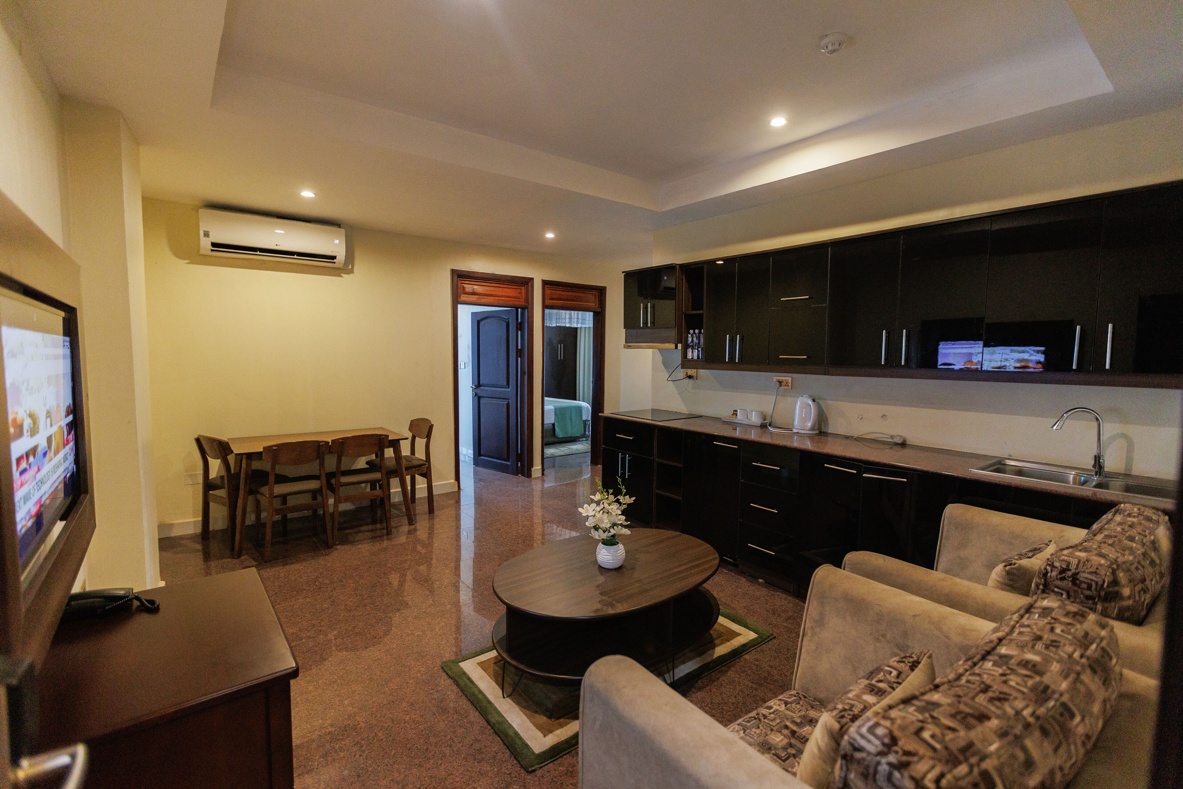 Una sala de estar moderna con un televisor de pantalla plana, un comedor con muebles de madera, una cocina con gabinetes oscuros y un lavamanos de acero inoxidable, y un pasillo que conduce a otra habitación.