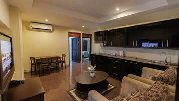 Una sala de estar moderna con un televisor de pantalla plana, un comedor con muebles de madera, una cocina con gabinetes oscuros y un lavamanos de acero inoxidable, y un pasillo que conduce a otra habitación.
