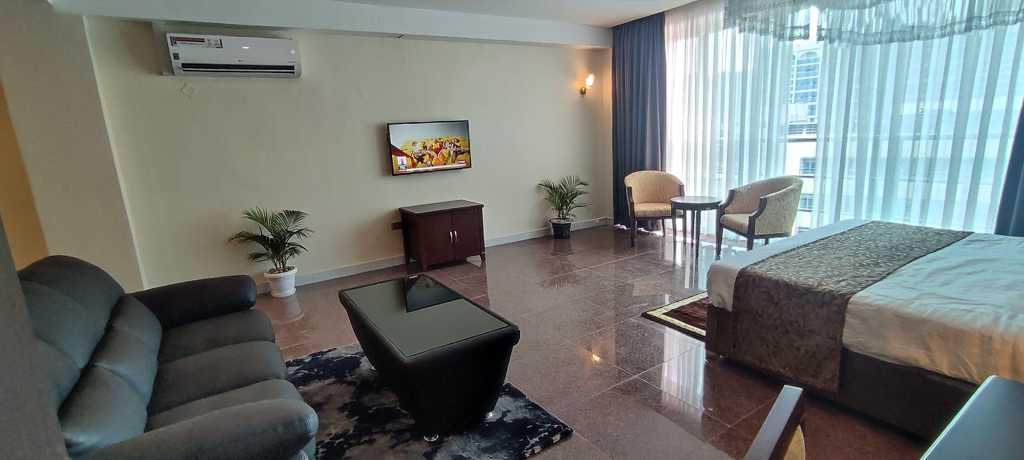 A hotel room with a bed, two armchairs, a small table, a TV, a cabinet, and two potted plants.