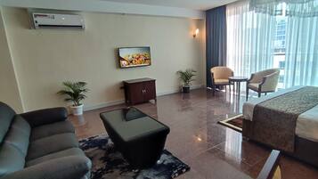 A hotel room with a bed, two armchairs, a small table, a TV, a cabinet, and two potted plants.