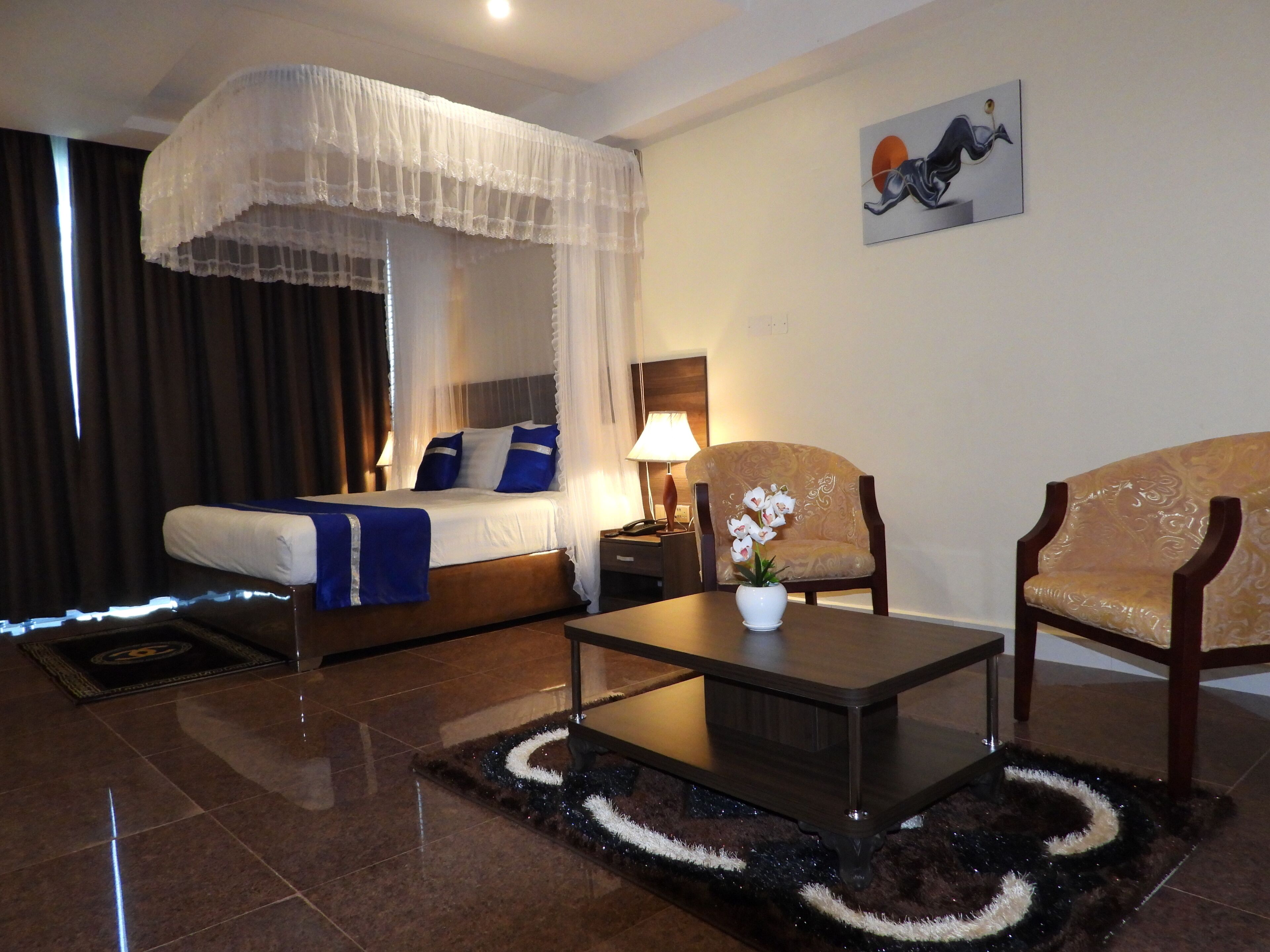 Habitación de hotel con una cama, dos sillones, una mesita de centro, una mesita de noche, una lámpara y un cuadro en la pared.