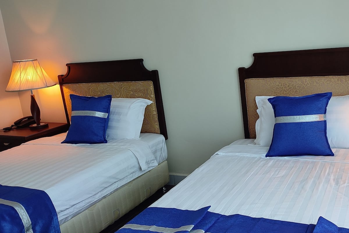 Dos camas individuales con sábanas blancas y almohadas azules, un cabecero de madera, lámparas de noche y una mesita de noche con teléfono.