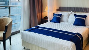 Une chambre d’hôtel avec un grand lit, une chaise, une table de chevet et une vue sur la ville grâce à de grandes fenêtres.