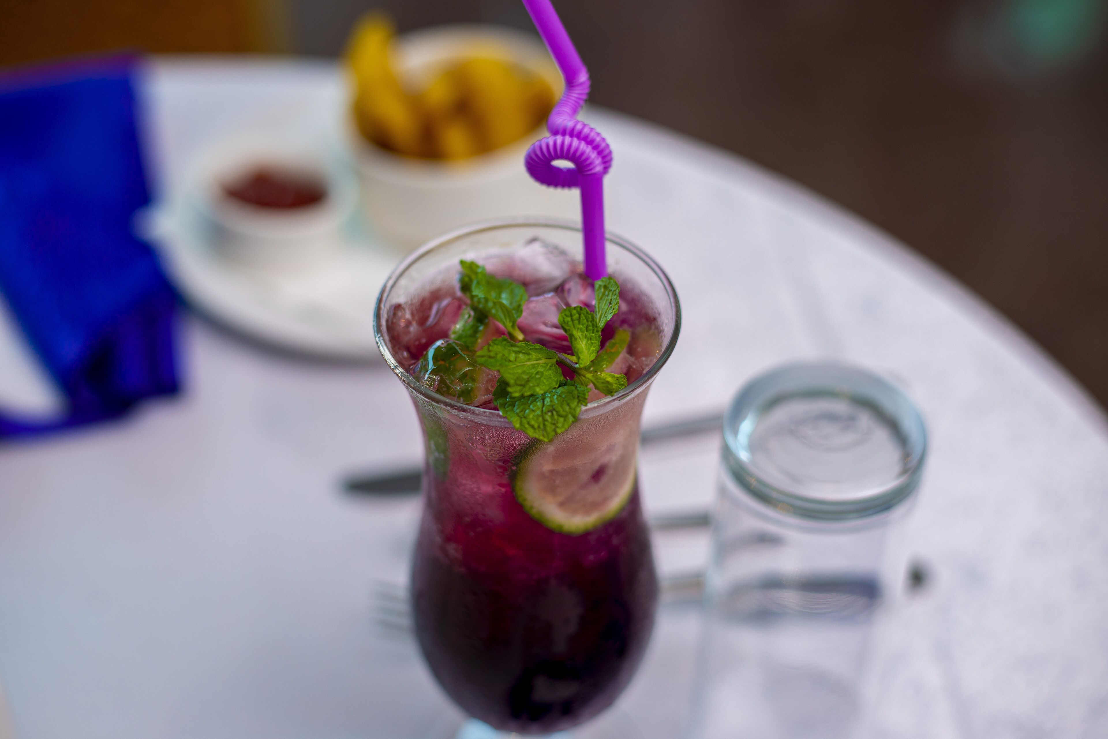 A glass of iced beverage with mint leaves and a lime slice, a small bowl of fruit, and a glass of water on a white table.
