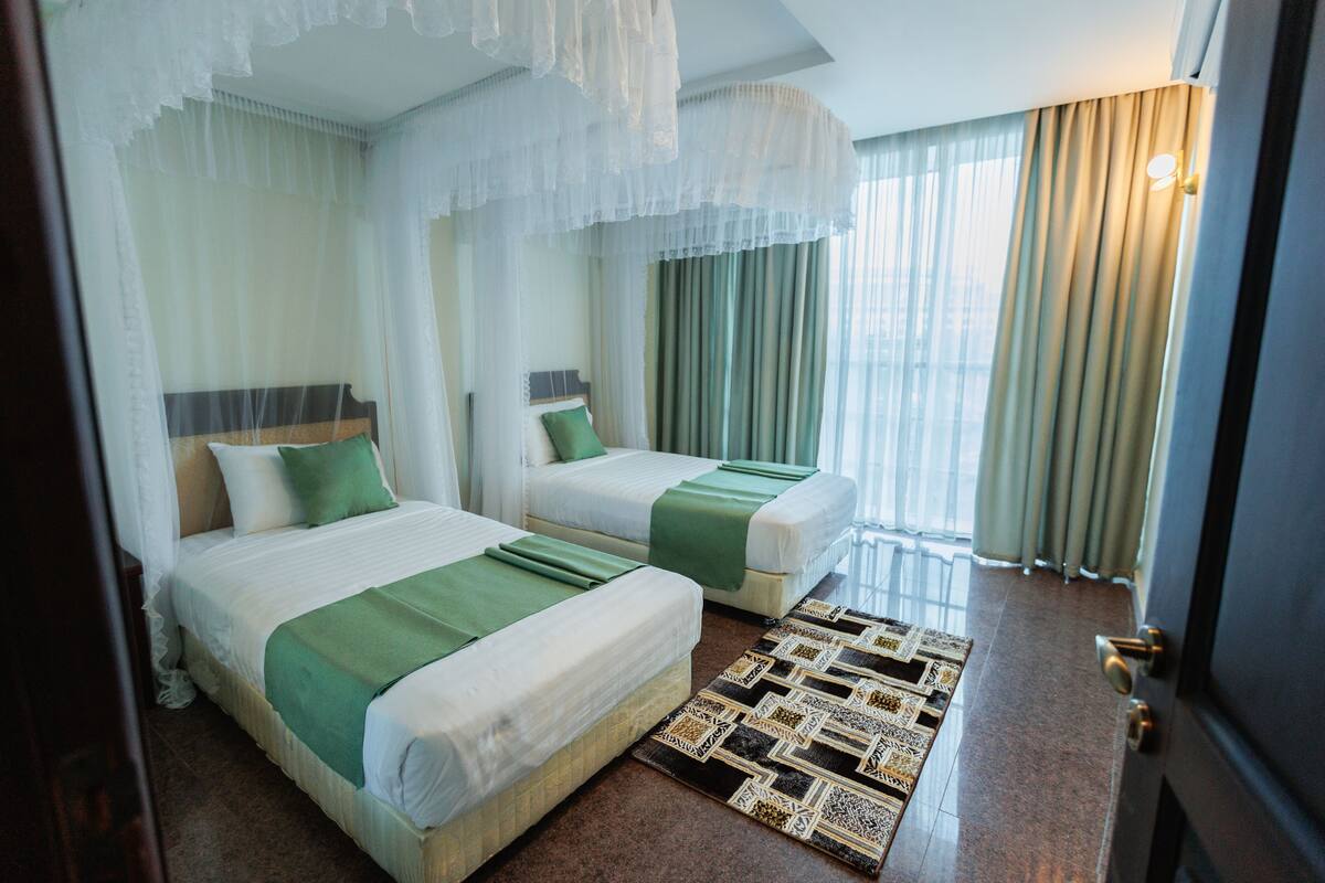 Habitación de hotel con dos camas, un tapete estampado y cortinas traslúcidas.