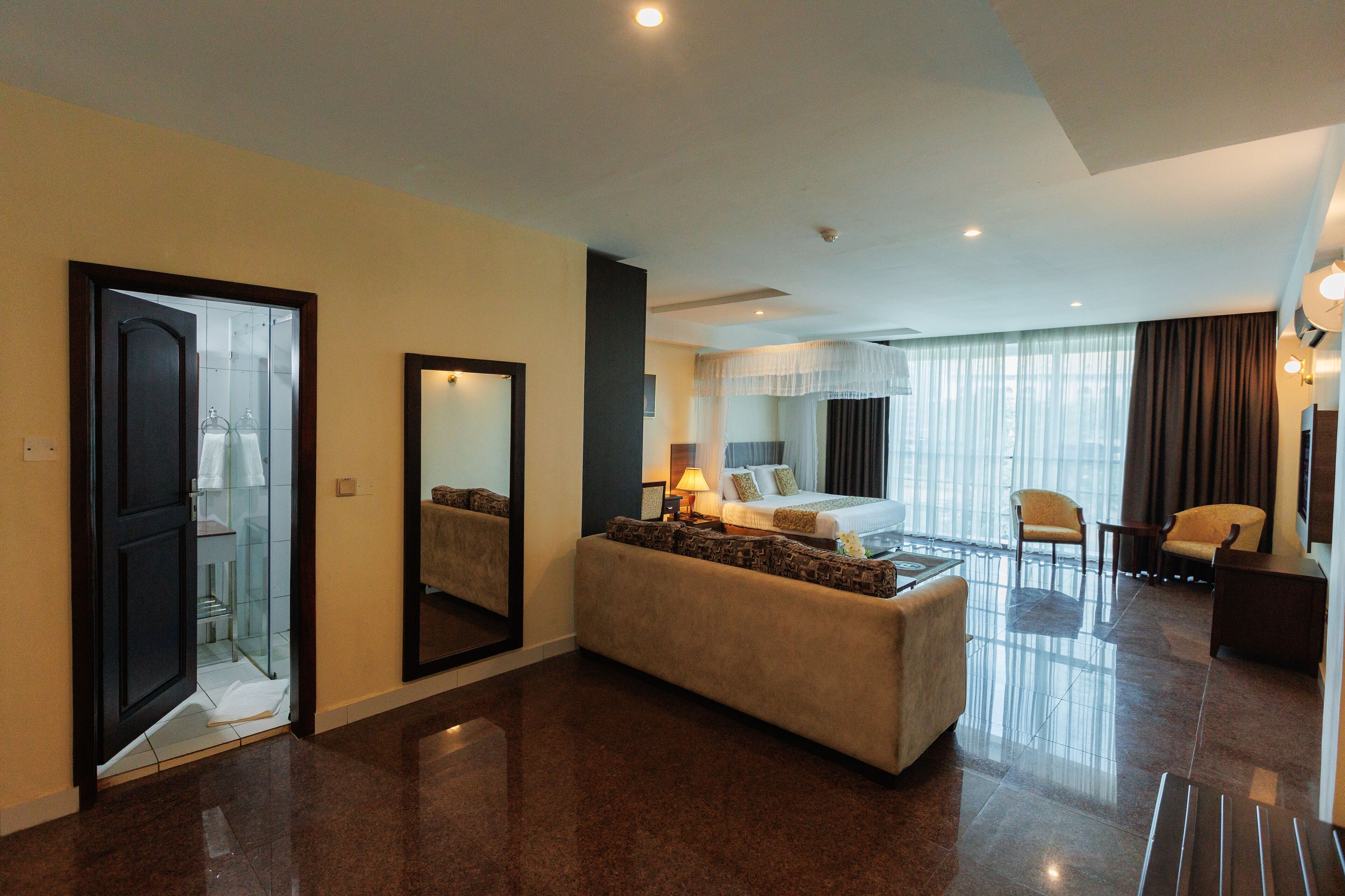 A hotel room with a large bed, a sitting area with a sofa, a mirror, and a view of the city through the window.