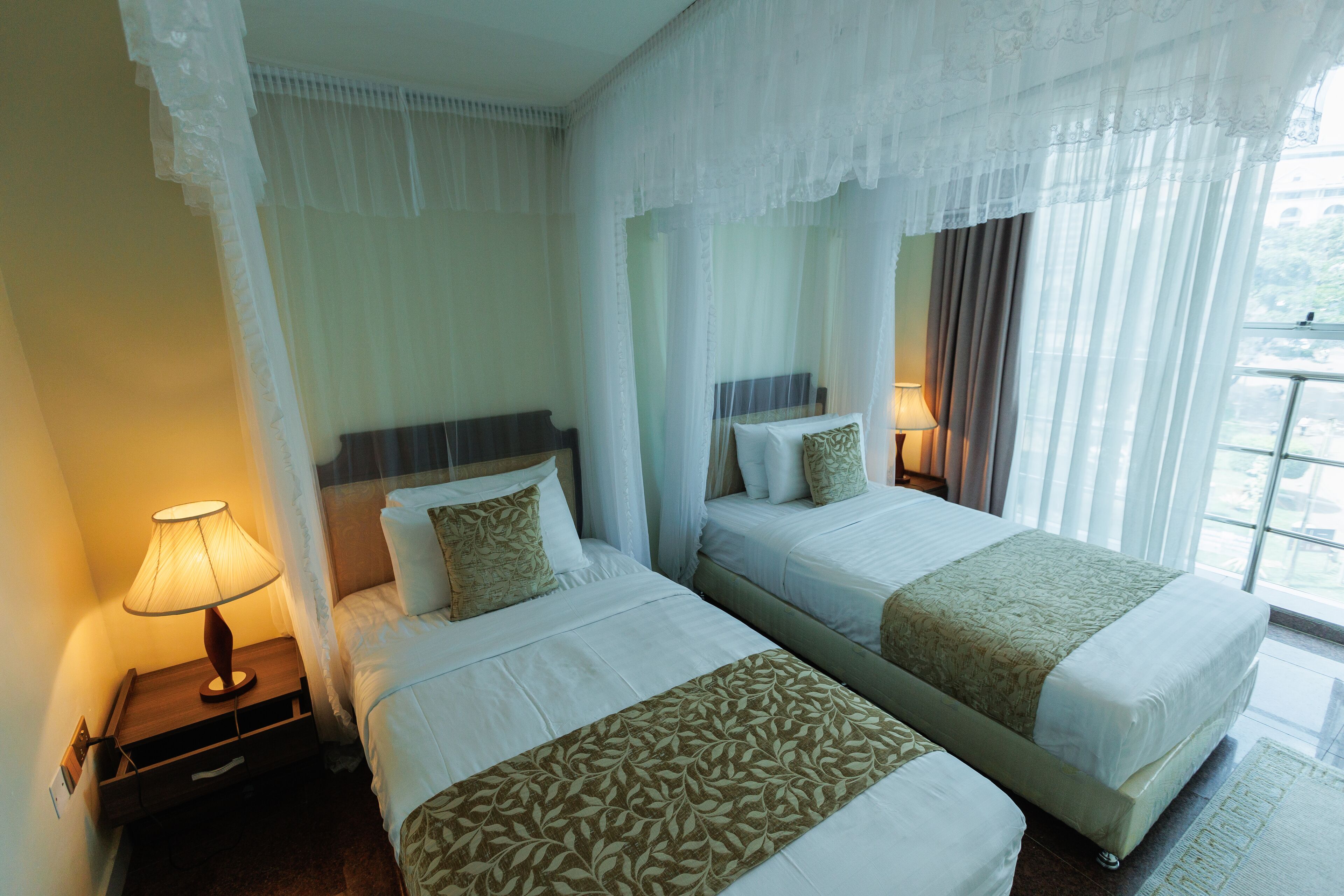 Une chambre d’hôtel avec deux lits, une table de chevet avec une lampe et des rideaux légers.